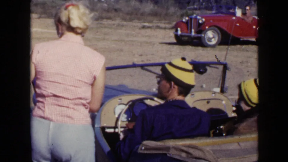 The woman stands beside the blue convertible while the seated driver looks downward, with another capped passenger partly visible.
