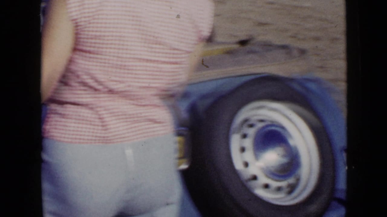 Close view of a blue car wheel and rear bodywork beside a person in a pink gingham top.