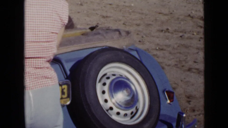A person in pink gingham stands beside the rear wheel of a blue convertible in the supplied 1949 San Diego home-movie footage.