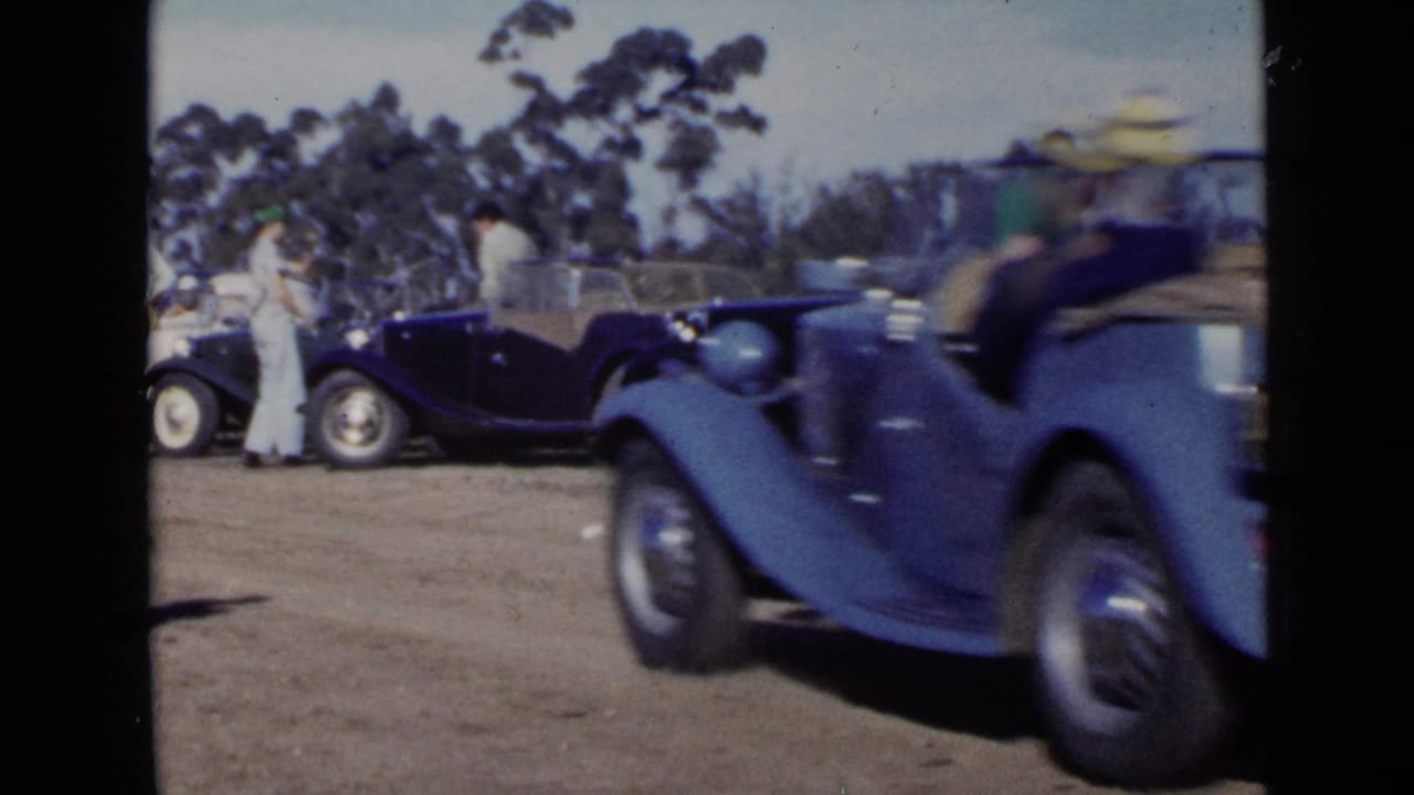 In 1949 San Diego home-movie footage, a blue convertible passes parked classic cars while adults stand nearby on a dirt lot.