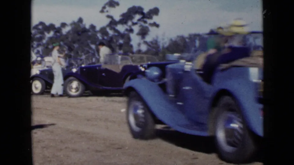 In 1949 San Diego home-movie footage, a blue convertible passes parked classic cars while adults stand nearby on a dirt lot.