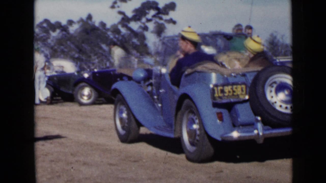 Rear view of a blue convertible with seated adults near parked classic cars.