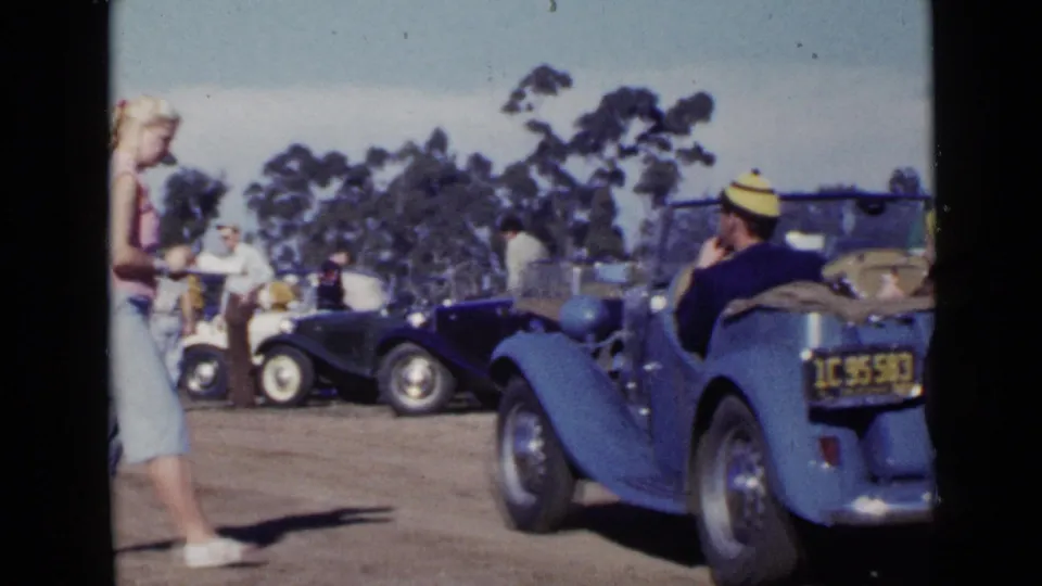 A woman in a pink top and light capri pants walks through the foreground as adults and classic cars fill the San Diego dirt-lot scene.