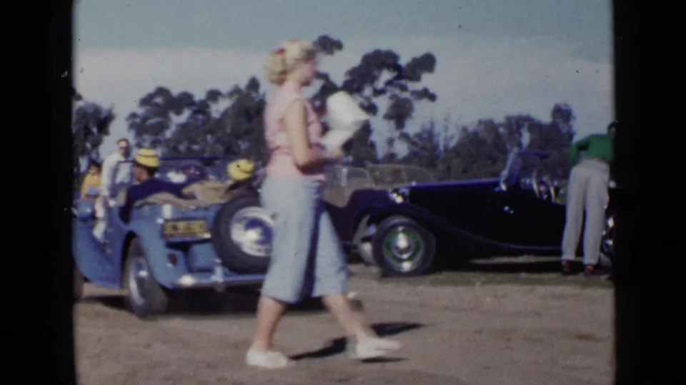 The foreground woman passes between the camera and a blue convertible, with dark classic cars and visitors visible in the 1949 San Diego scene.