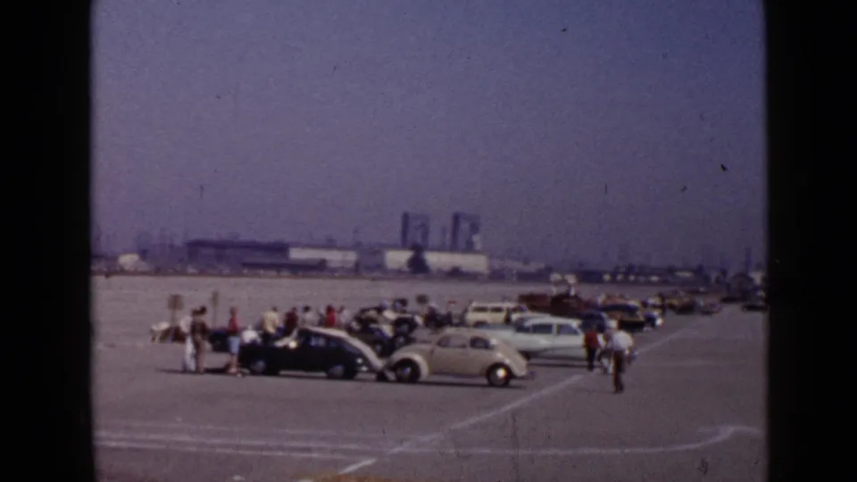 Distant 8mm view of a race-pit or parking area in the supplied Southern California location, with spectators, parked cars, and industrial buildings.