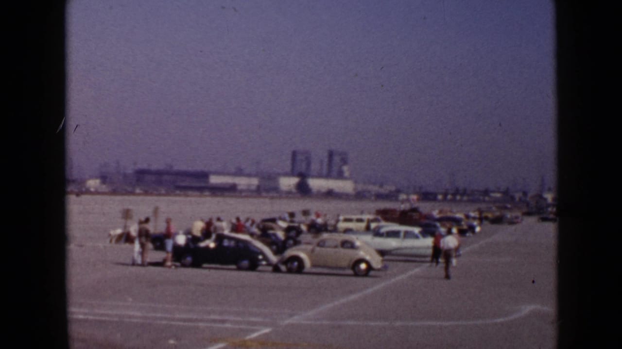 Wide view of vintage cars and spectators on a large paved lot with industrial buildings in the distance.
