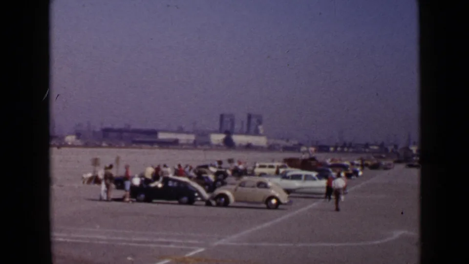 1949 San Diego home-movie frame showing people gathered around parked vintage cars on a broad paved lot near distant industrial structures.