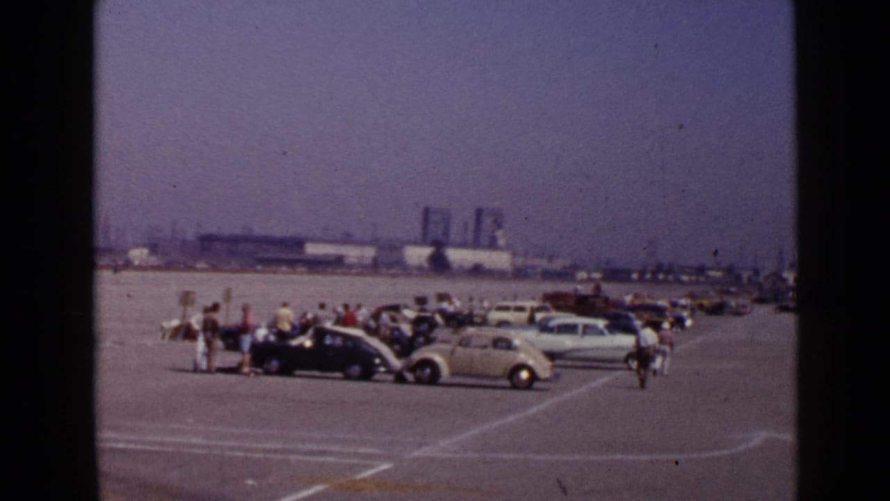 1949 San Diego frame with visitors clustered near vintage automobiles on an open lot, with low industrial buildings in the background.
