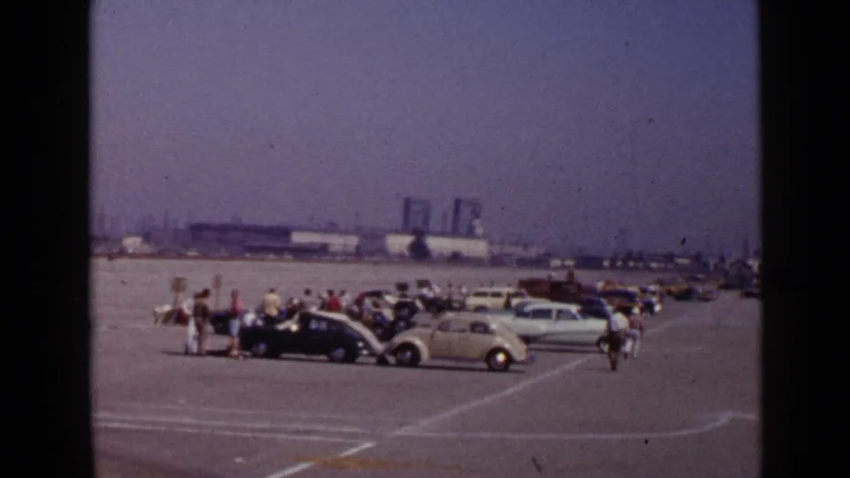 1949 San Diego frame with visitors clustered near vintage automobiles on an open lot, with low industrial buildings in the background.