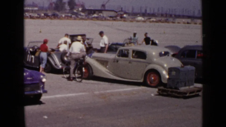 Closer 1949 San Diego home-movie view of adults gathered around older cars, with a bicyclist beside a long cream sedan on the paved lot.