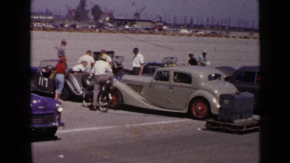 1949 San Diego frame showing visitors around a cream-colored sedan and dark vintage cars, with distant vehicles and industrial structures behind them.