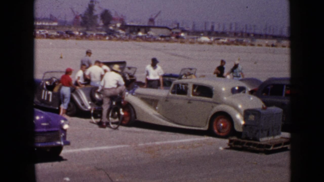 Men gather around classic cars in a sunny paved lot with cranes in the distance.