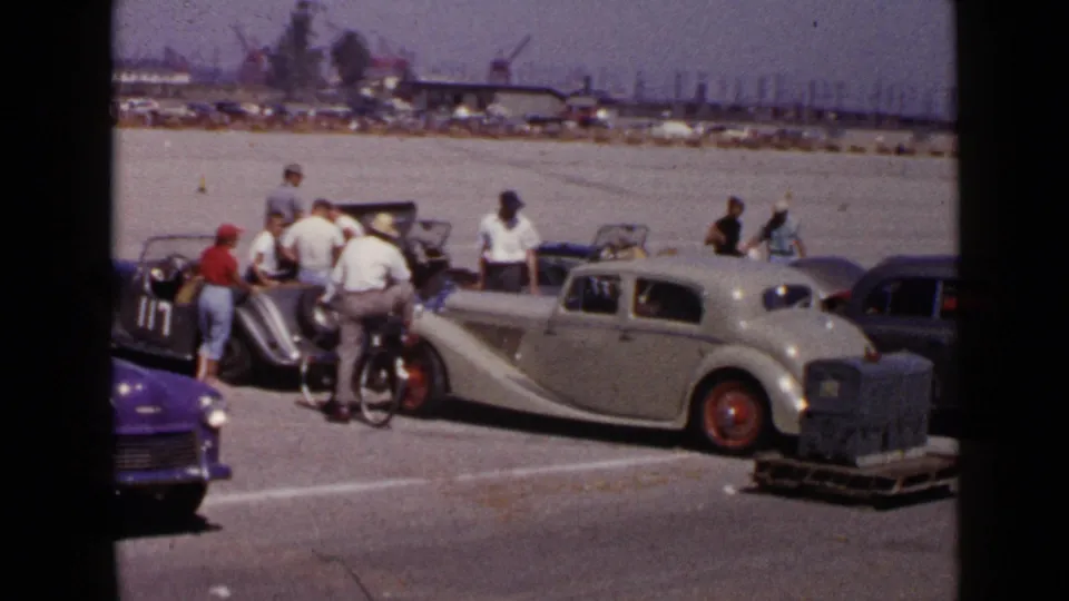 1949 San Diego home-movie frame showing men around a silver sedan and other classic cars in a bright open lot near distant cranes.