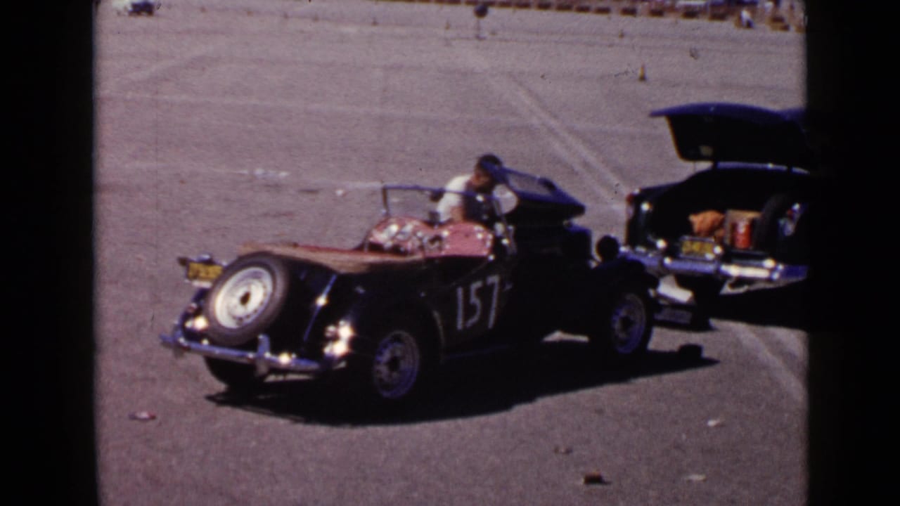 Black vintage open-top car with number 157 is parked in a wide paved area beside another car with an open trunk.