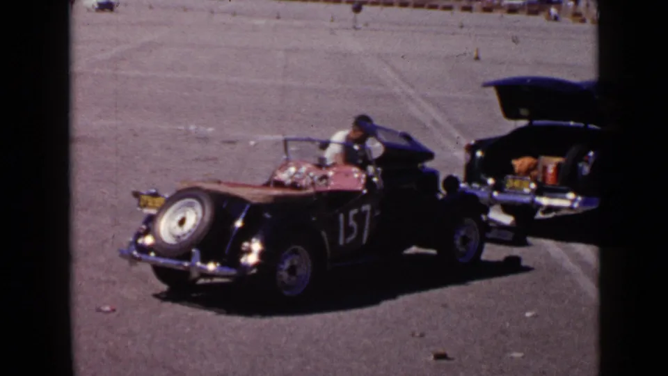 Black vintage open-top car with number 157 is parked in a wide paved area beside another car with an open trunk.