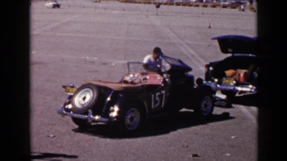 1949 San Diego frame showing the black number 157 vintage car angled across the lot next to a parked car with its trunk open.