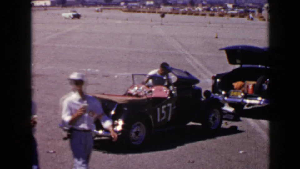 A man in a light cap walks through the foreground as the black number 157 vintage car remains parked in the San Diego lot.