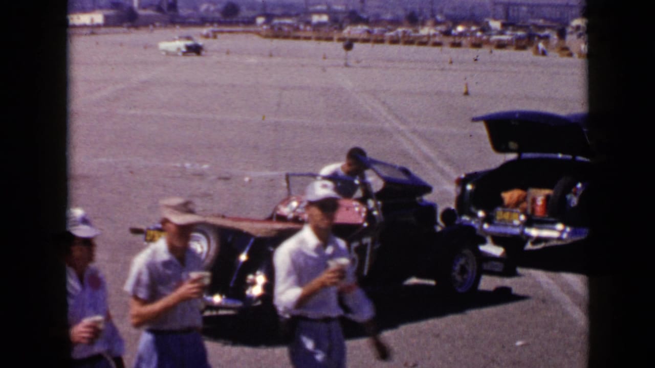 1949 San Diego home-movie frame showing men near a black open-top race car numbered 157 with another car trunk open nearby.