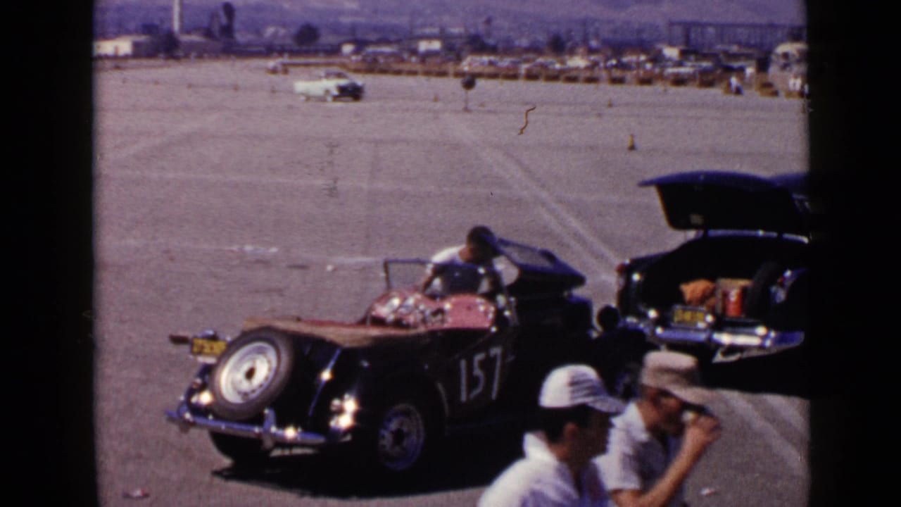 Race car 157 sits beside an open trunk as men pass in the foreground.