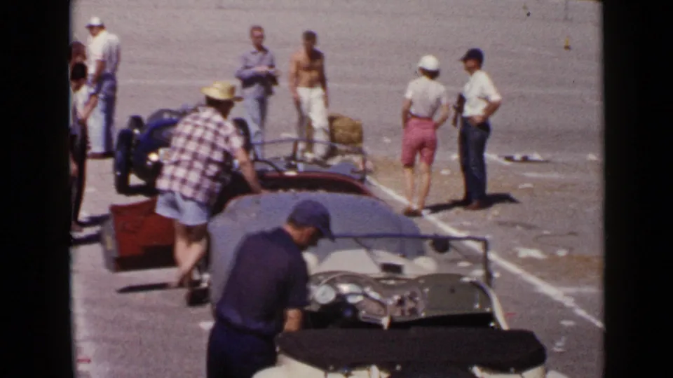 1949 San Diego home-movie frame showing men gathered around open race cars, including a foreground cockpit and a small blue roadster.