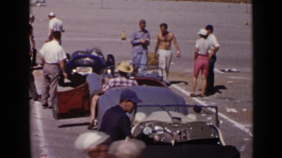 1949 San Diego frame showing a foreground roadster cockpit, men in caps, and another small race car in a paved preparation area.