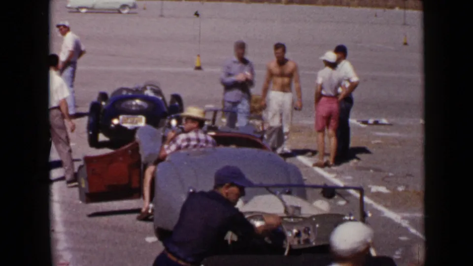 San Diego 1949 home-movie frame showing men gathered near open-top sports cars on a paved lot.