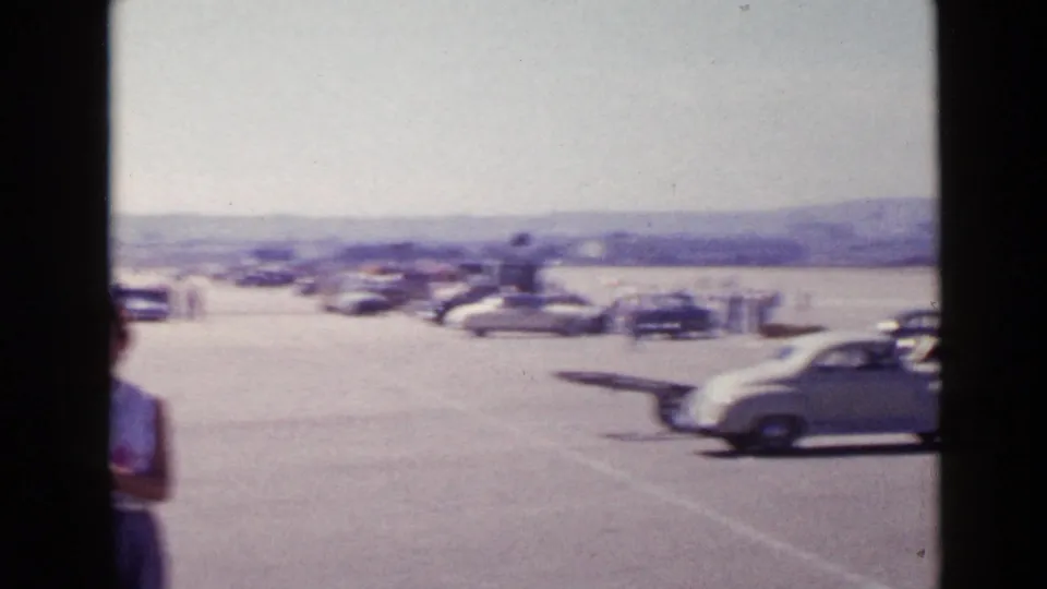 Soft 8mm frame from the San Diego gathering, with cars crossing a broad paved area below distant hills.