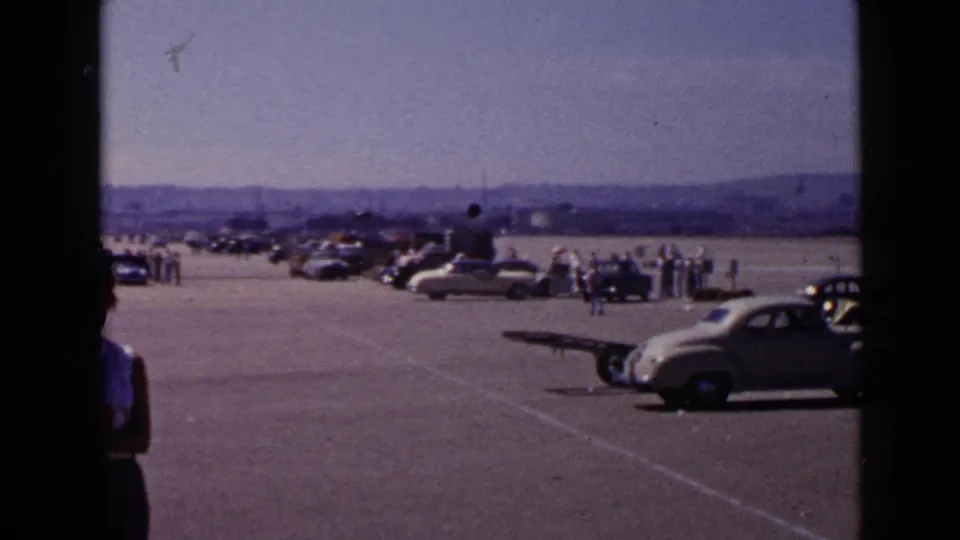 San Diego 1949 wide home-movie view of cars and people spread across a paved gathering area under distant hills.