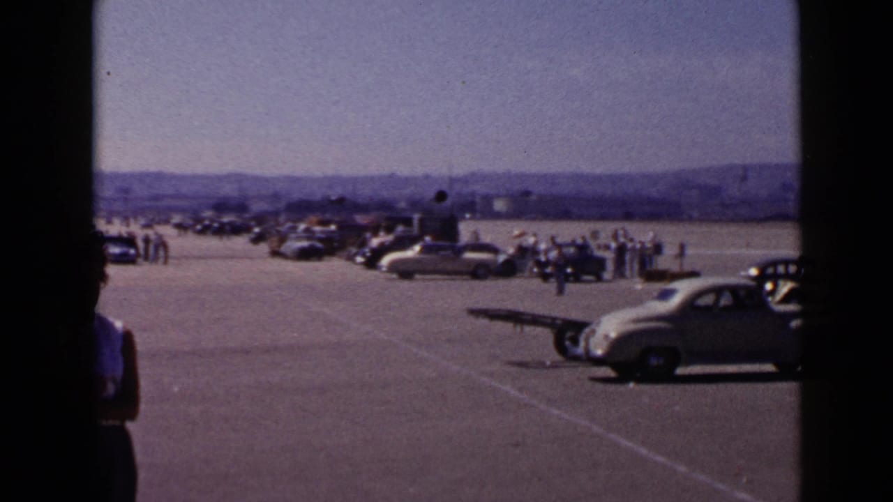 Parked vintage cars and small groups of people on a broad paved airfield.