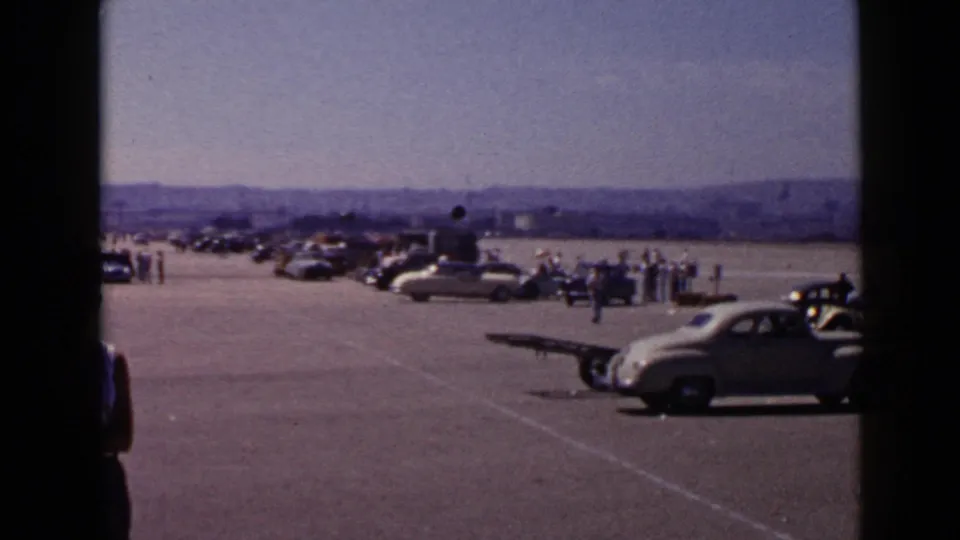1949 San Diego footage frame with vintage automobiles, scattered spectators, distant low hills, and a partially obstructed view.