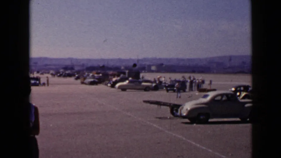 Home-movie view of a San Diego airfield gathering with period cars, people in light clothing, and distant hills in 1949.
