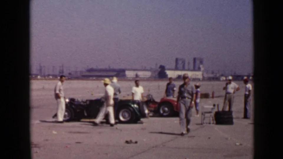 1949 San Diego frame showing people near a small open car, with industrial-looking structures in the distant background.