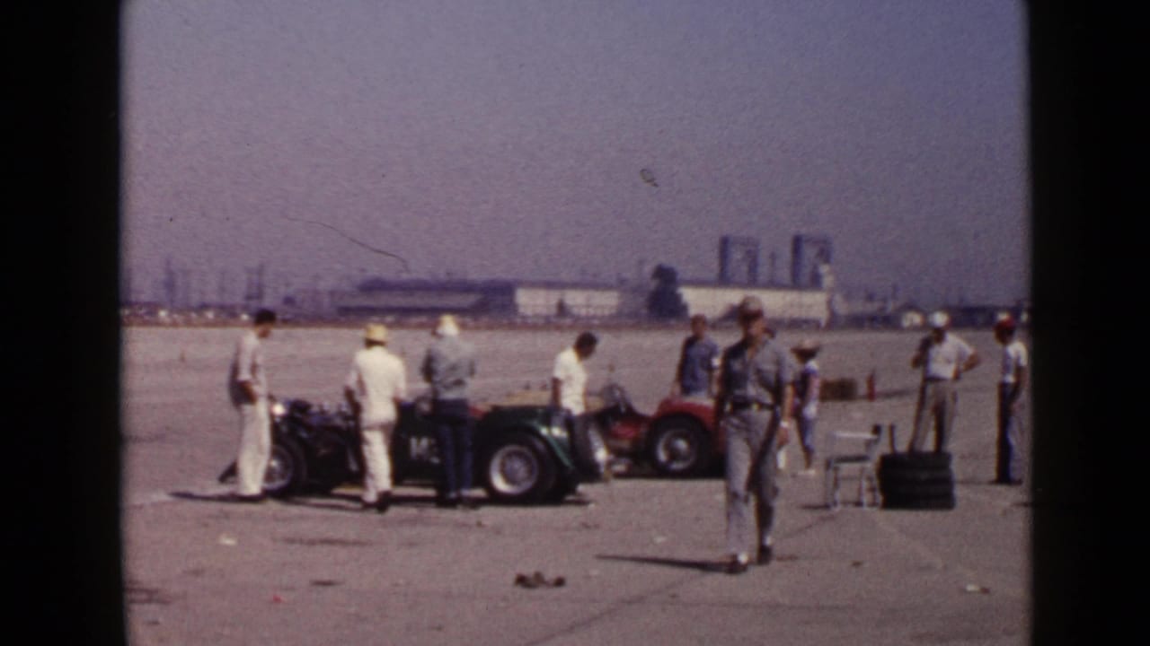 Men stand around vintage open cars on a broad paved airfield with industrial buildings in the distance.