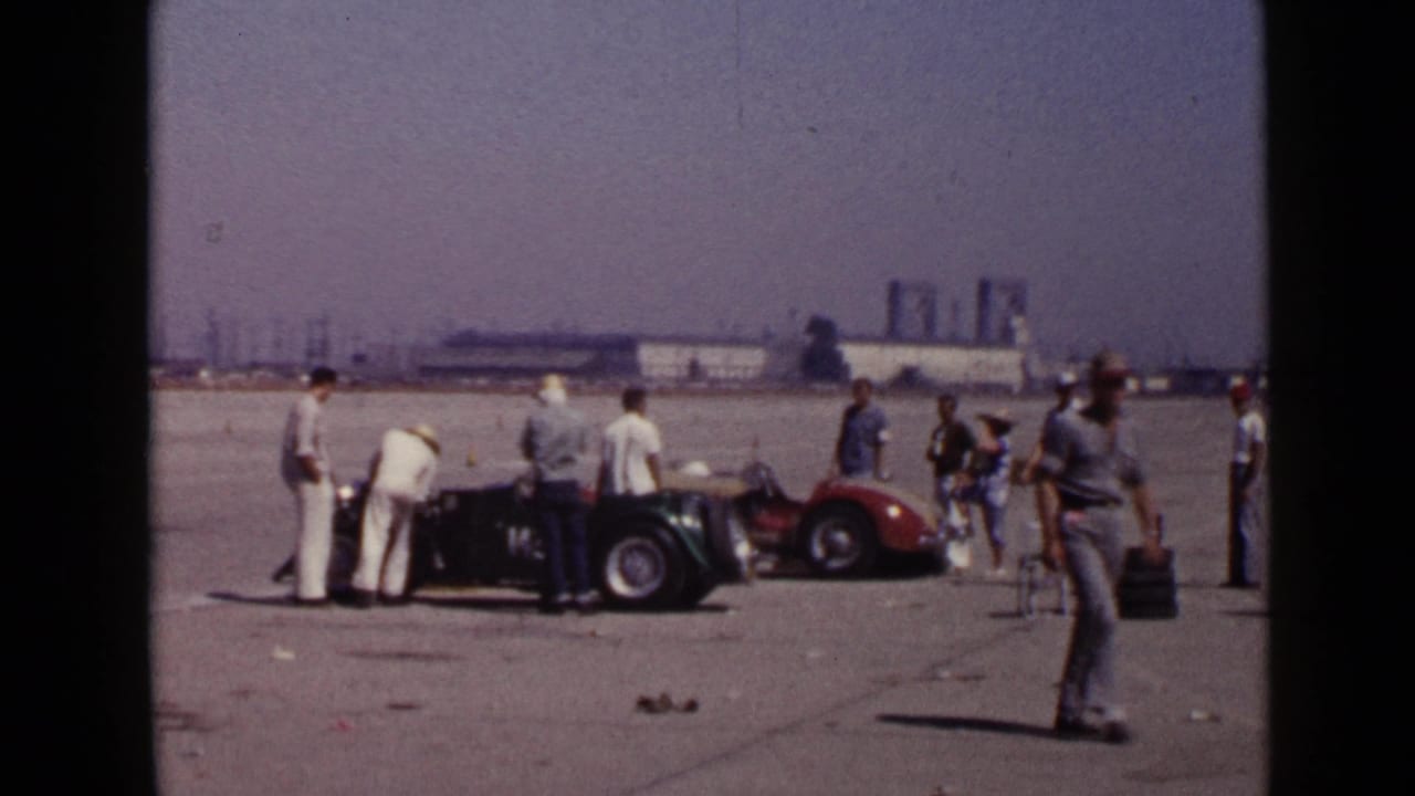 A 1949 San Diego frame shows men gathered around vintage open cars as one man walks across the foreground of the airfield setting.
