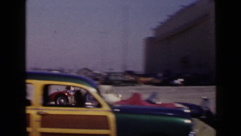 A colorful station wagon, race cars, and several people appear near the San Diego racetrack pit area in 1949 footage.