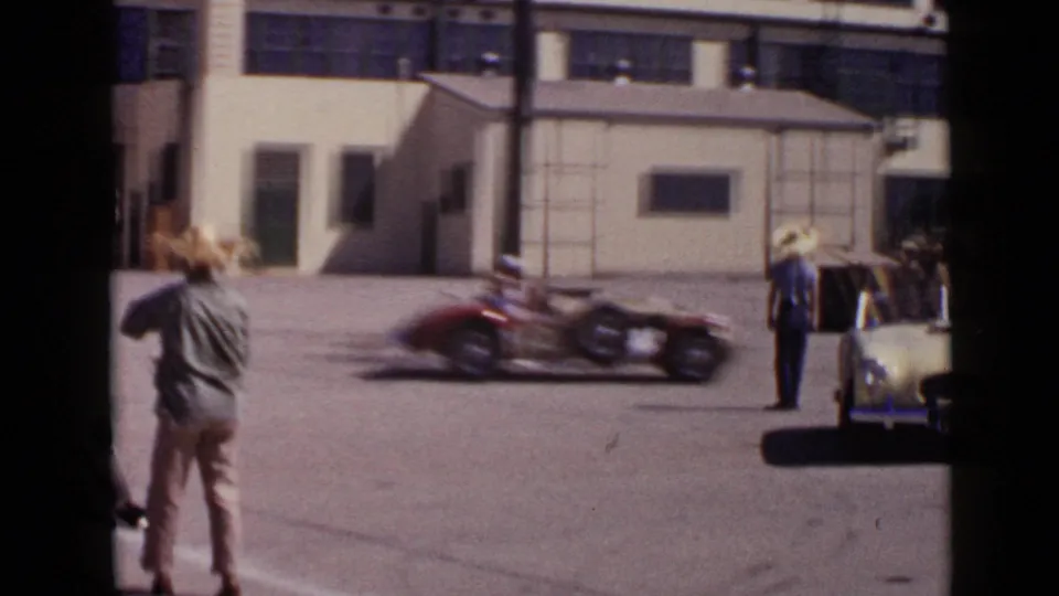 A dark open-wheel race car moves quickly past industrial buildings and onlookers in 1949 San Diego racing footage.
