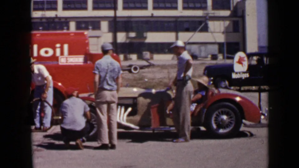 Pit crew and spectators stand around a vintage race car near service vehicles and visible signage in 1949 San Diego footage.