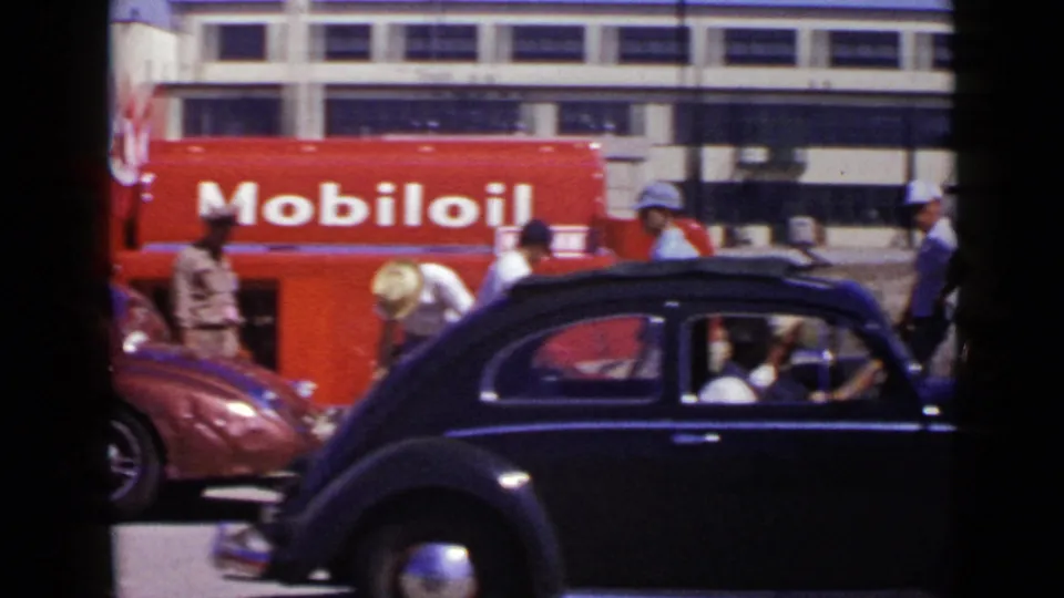 1949 San Diego frame where a black vintage car crosses the foreground as men and a red convertible remain near the Mobil Oil truck.