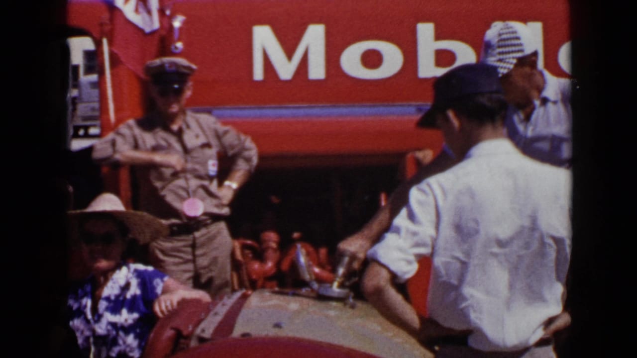 Uniformed man standing by a red Mobil truck as spectators gather near a decorated car.