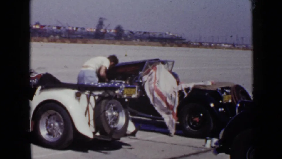 1949 San Diego home-movie frame showing a man working at an open decorated convertible with flag-patterned fabric at the side.