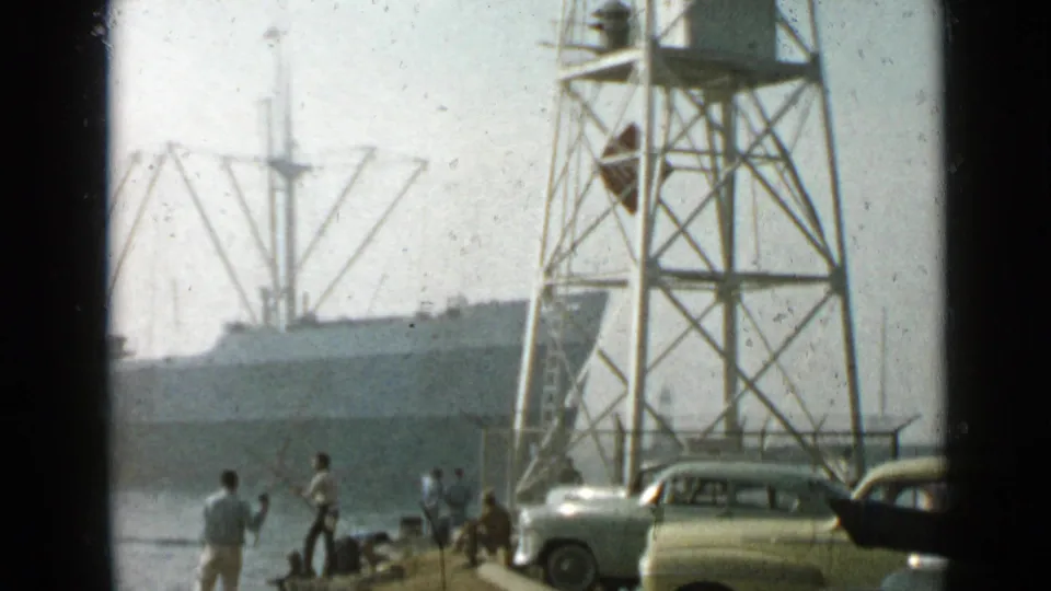 Long Beach waterfront scene with a metal tower in the foreground, parked period cars, fishermen on the pier, cranes, and a cargo ship beyond.