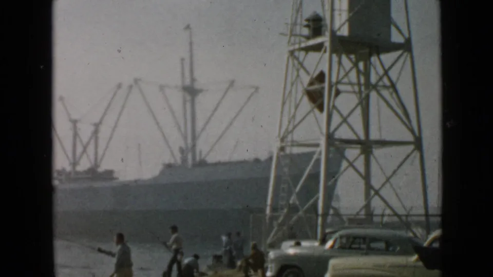 Waterfront view at Pierpoint Landing showing people near the pier, a metal tower, parked cars, harbor cranes, and a cargo ship in hazy light.