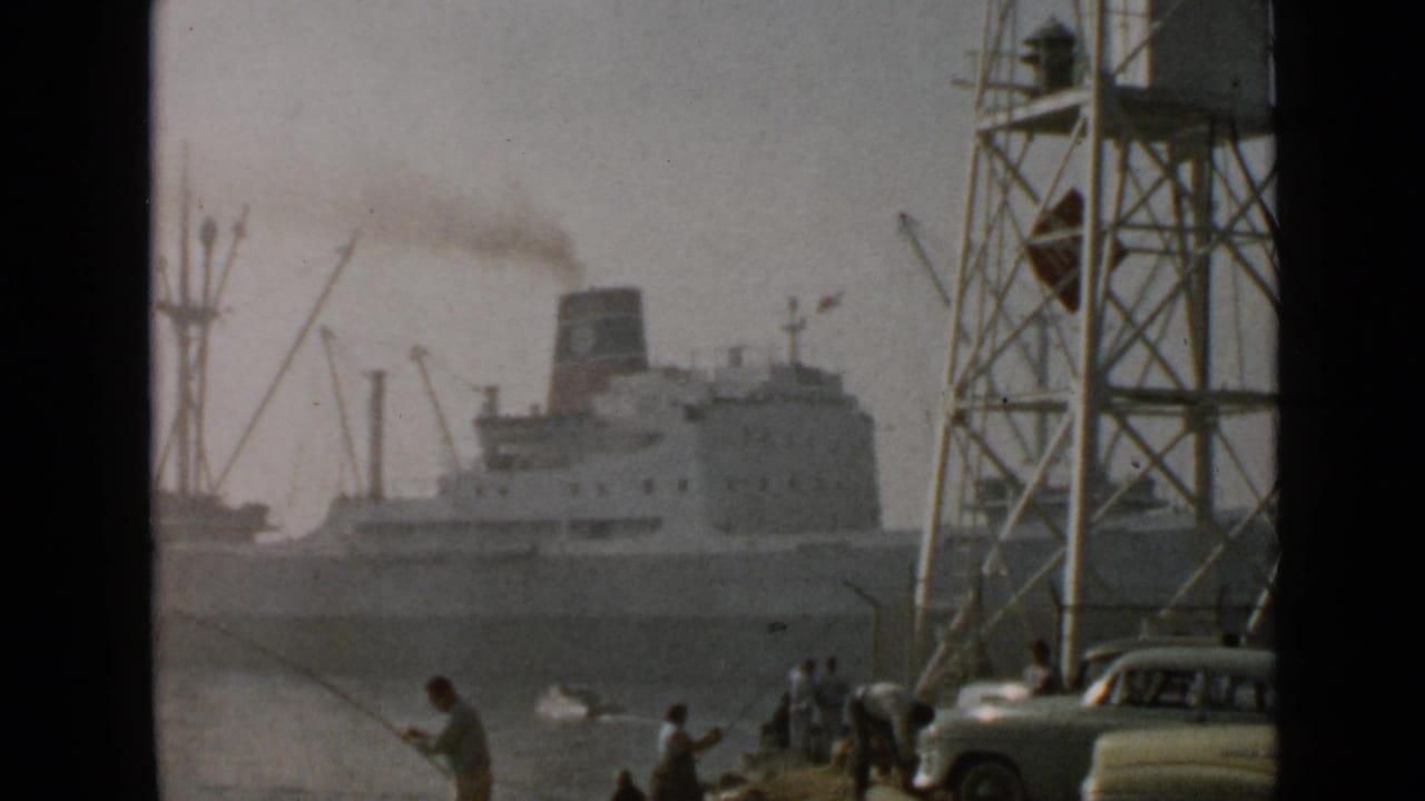 1949 San Diego harbor view with a smoking steamship, dock cranes, shoreline fishermen, a metal tower, and a mid-century car.