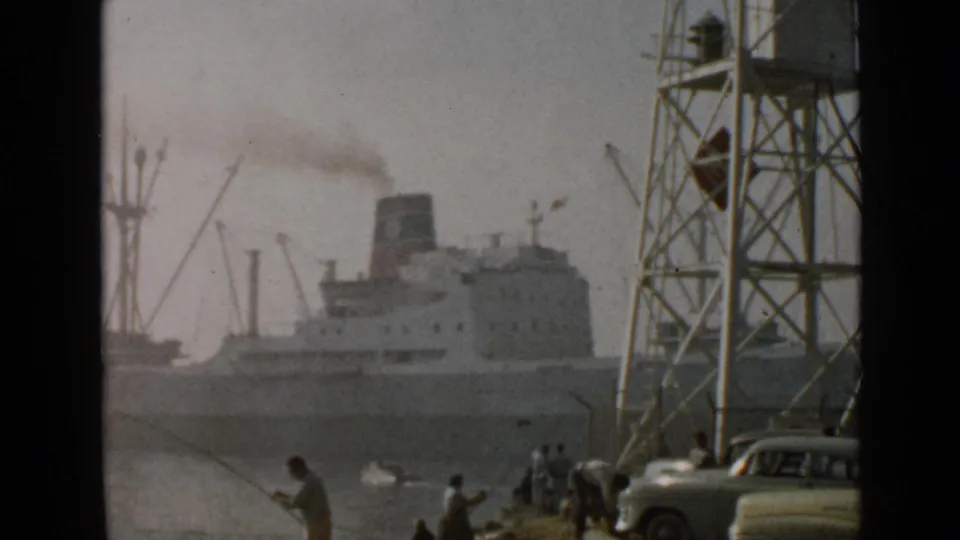 1949 San Diego harbor view with a smoking steamship, dock cranes, shoreline fishermen, a metal tower, and a mid-century car.