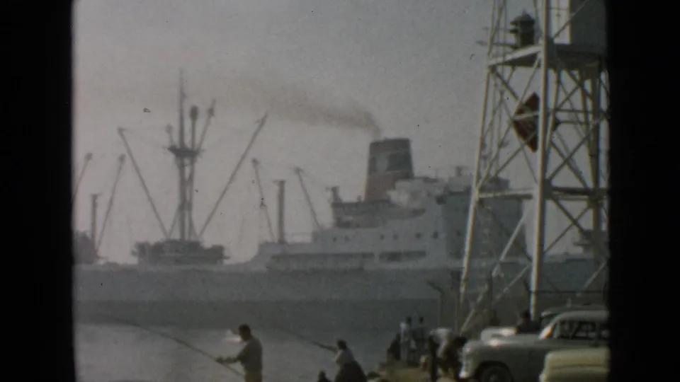 Soft 8mm frame of a 1949 San Diego harbor, showing a smoking ship, cargo cranes, fishermen, and a car partly visible at the edge.