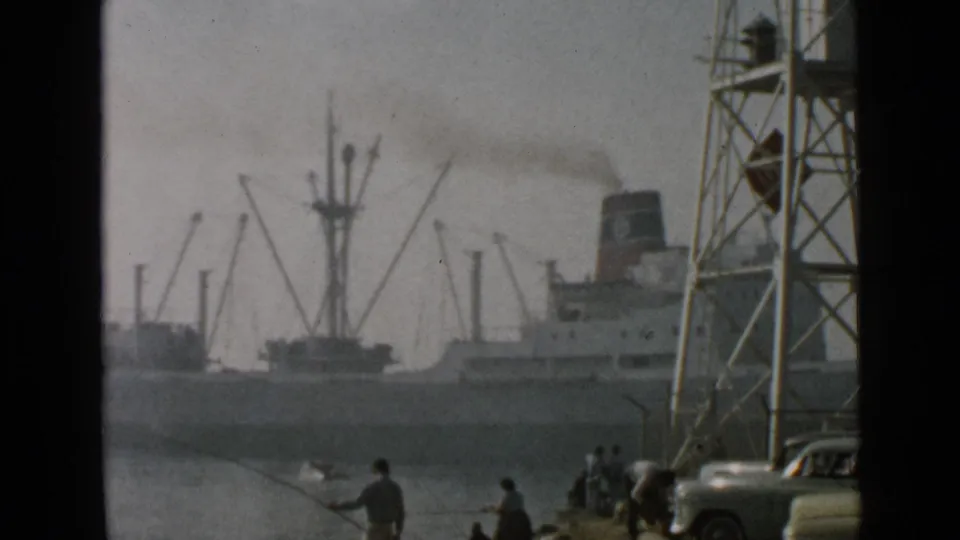 1949 San Diego port view with a large smoking ship, industrial cranes, a foreground tower, fishermen, and a classic car near the shore.