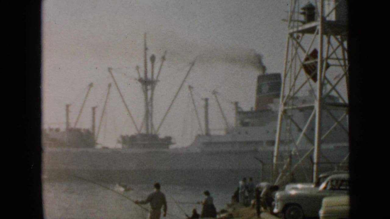 Large steam cargo ship passing fishermen near parked classic cars and dock cranes.