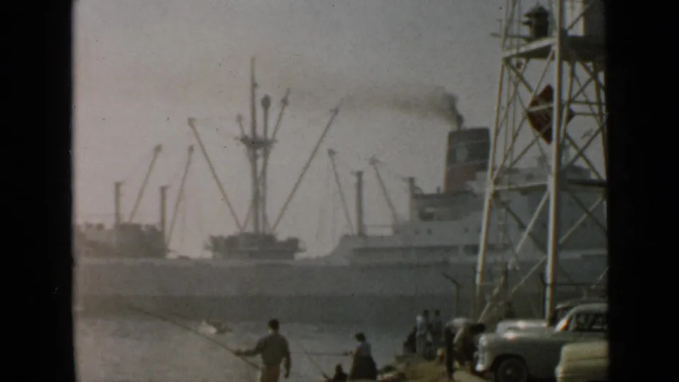 1949 San Diego harbor view with a smoking cargo ship moving behind shoreline fishermen, parked cars, and port cranes.