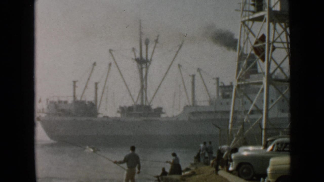 1949 San Diego harbor scene with a smoking cargo vessel, cranes, shoreline anglers, and classic automobiles at the water's edge.