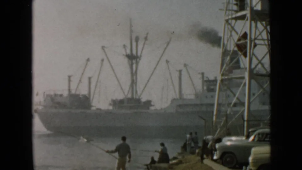 1949 San Diego harbor scene with a smoking cargo vessel, cranes, shoreline anglers, and classic automobiles at the water's edge.